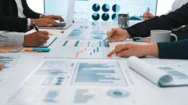 Bir grup profesyonel, modern ofis ortamında proje kararlarını bilgilendirmek için veri şemaları ve grafikleri analiz ederek canlı bir iş toplantısı düzenliyor. SACTR