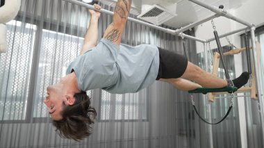Çekirdek dayanıklılığı ve spor salonunda vücut egzersizi için pilates trapez masasına tırmanan bir adam. Eğitmen, arka ve kalça esnekliğini arttırmak için Cadillac 'ta ters asılı duruyor. Alışkanlık.
