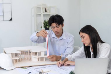 Profesyonel Asyalı erkek mimar ev modelinin uzunluğunu ölçmek için cetvel kullanırken, beyaz tenli genç meslektaşı ev modeliyle toplantı masasındaki verileri analiz etmek için dizüstü bilgisayar kullanıyor. Tertemiz..