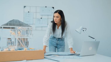 Genç, güzel beyaz bir mimar masanın üzerine plan koyar ve ev modeliyle karşılaştırırken, modern ofisteki fahişe modellerini dikkatlice incelerken arkadaki çizgilerle karşılaştırır. Tertemiz..
