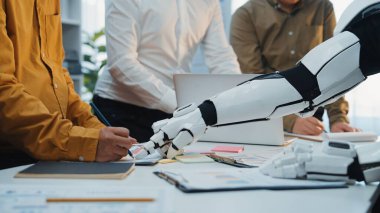 Bir insan ve bir robot el modern ofis ortamında işbirliği yapıyor. Bu sahne gelecekteki takım çalışmasındaki teknoloji ve yenilik sinerjisini temsil ediyor. Noogenesis