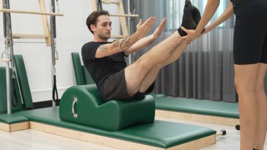 Erkek müşteri pilates ark fıçısında bacak kaldırma alıştırması yapıyor. Vücut dengesini, bacak dayanıklılığını ve spor salonundaki esnekliğini arttırmak için eğitmen yardımı ile birlikte. Öğrenci yalancı ve muziplik pozu veriyor. Alışkanlık.