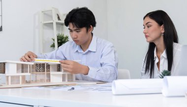 Profesyonel Asyalı erkek mimar ev modelinin uzunluğunu ölçmek için cetvel kullanırken, beyaz tenli genç meslektaşı ev modeliyle toplantı masasındaki verileri analiz etmek için dizüstü bilgisayar kullanıyor. Tertemiz..