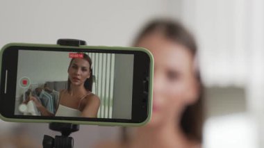 Genç sosyal medyum kadın moda videosu yapıyor. Blogcu kameraya selfie çubuğu ve ışık halkası kullanarak gülümserken, aynı zamanda ikna edici çevrimiçi kıyafetlerin izleyicilere veya takipçilere vlog satmasını sağlıyor. Düzenle