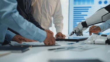 Şık bir ofis ortamında dijital tablet üzerinde insan eliyle robot kol arasındaki işbirliğini gösteren modern bir iş toplantısı. Noogenesis