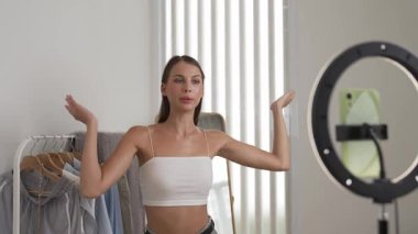 Genç sosyal medyum kadın moda videosu yapıyor. Blogcu kameraya selfie çubuğu ve ışık halkası kullanarak gülümserken, aynı zamanda ikna edici çevrimiçi kıyafetlerin izleyicilere veya takipçilere vlog satmasını sağlıyor. Düzenle