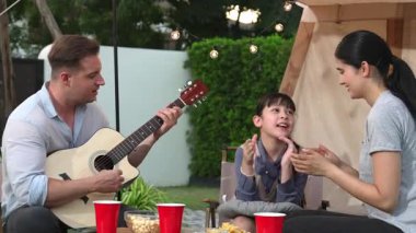 Baba gitar çalarken anne ve kız birlikte kampta oturuyorlar. Ebeveynler, genç nesil çapraz kuşak farklılıklarıyla zaman geçirmek için açık hava kamp aktivitelerini kullanıyorlar. Uyumsuzluk.