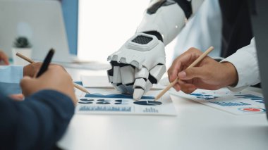 Bir insan eli ve bir robot el, günümüz dijital dünyasında takım çalışması ve teknolojik yeniliklerin altını çizerek, modern ofis alanındaki veri raporlarını işbirliği içinde analiz eder.