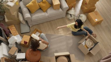 Oturma odasındaki mobilyaların taşınmasına yardım ederken yerde oturan mutlu oda arkadaşı. Yeni eve taşınmak için birlikte çalışan Asyalı lezbiyen aşığının havadan görünüşü. Yer değiştirme konsepti. Myrmidon.