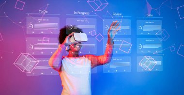 Modern bir ofiste dijital panolar ve etkileşimli elementlerle çevrili, görev yönetimi ve çevik proje planlaması için sanal gerçeklik teknolojisiyle uğraşan genç bir profesyonel. XDMCP