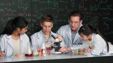 Çok kültürlü bir öğrenci mikroskop altında incelerken güzel bir kız da numuneyi inceliyor. BTMM sınıfında karatahta bilim teorisi deneyi yapan liseli çocuklara bakan öğretmen. Öğretim