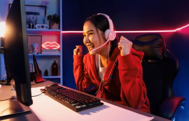 Yüzünde kulaklık renginde kulaklık takan, neon odada online oyun oynayan, Esport meziyetli takım oyuncularına yumruk kaldırarak oynayan güzel Asyalı kız flamasının sunucu kanalı. Taktik.