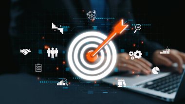Bu resim, iş simgeleri ve grafiklerle çevrelenmiş, modern bir çalışma alanındaki hedef başarısını ve stratejik planlamayı sembolize eden, bir okun merkeze isabet ettiği dijital bir hedefe sahiptir. Dörtgen