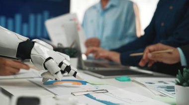 Robot bir el, canlı bir iş toplantısında, teknolojinin ve insan işbirliğinin modern çalışma alanlarına entegrasyonunu gösteren veri grafiklerine uzanır. Noogenesis
