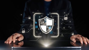 Güvenlik ve veri korumasını temsil eden grafiklerle çevrili bir dizüstü bilgisayarla uğraşan profesyonel, dijital bilginin korunmasının önemini vurguluyor. Dörtgen