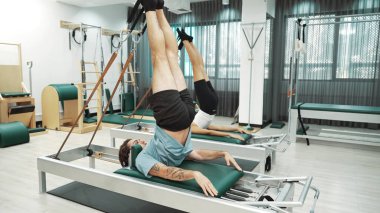 Müşteri, spor salonundaki yenilikçi bir makinede kayışla ters bacak kaldırma hareketi yapıyor. Çekirdek bağlantısını, dengesini ve esnekliğini geliştirmek için ters takla atan iki mutlu antrenör. Alışkanlık.