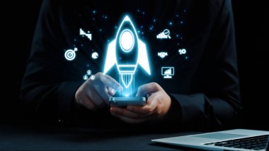 Bir iş adamı, parlayan roket fırlatma ikonlarıyla çevrili akıllı bir telefonla iletişim kurarak, modern iş ortamında yenilik, büyüme ve stratejik düşünmeyi sembolize eder. Dörtgen