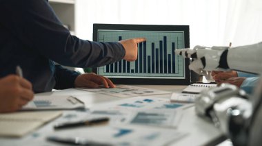 Modern bir iş ekibi veri analizi sunumuna girişiyor, gösterişli bir ofis ortamında robot bir kolla iş birliği yaparken ekranda bir tablo sergiliyor. Noogenesis