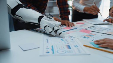 Veri analizi ve proje planlamasında takım çalışmasının vurgulandığı, iş toplantısı sırasında insan eliyle çalışan bir robot kolu gösteren dinamik bir sahne. Noogenesis