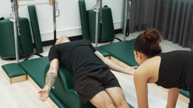Erkek müşteri pilates ark fıçısında bacak kaldırma alıştırması yapıyor. Vücut dengesini, bacak dayanıklılığını ve spor salonundaki esnekliğini arttırmak için eğitmen yardımı ile birlikte. Öğrenci yalancı ve muziplik pozu veriyor. Alışkanlık.