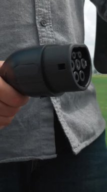 İlerici adam elektrik istasyonundan elektrik arabasına elektrik kablosu bağlıyor. Yeşil enerjiyle çalışan EV arabası gelecekteki temiz çevre konsepti için yeşil alanda çalışıyor..