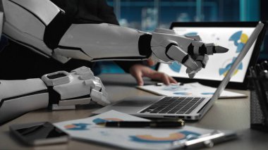 Robot bir el, modern bir ofiste teknoloji ve insan çabası arasındaki sinerjiyi sembolize eden iş analizlerini gösteren bir dizüstü bilgisayarla etkileşime girer. Noogenesis