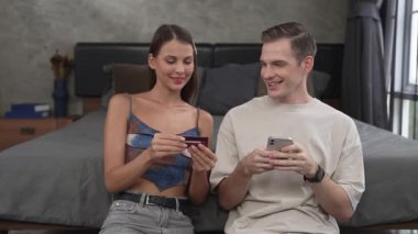 Genç çift, kredi kartıyla ödemek için internet üzerinden ödeme uygulaması ve dijital cüzdan kullanarak yatak odasında oturuyor. Mobil internet üzerinden alışveriş ve modern alımlar. Düzenle