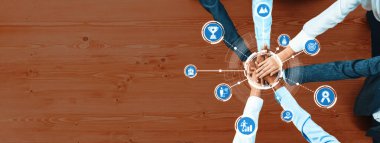 Ahşap zemine karşı başarıyı, hedefleri ve ortaklığı sembolize eden simgelerle çevrelenmiş el ele tutuşan takım çalışması ve işbirliğinin görsel bir temsili. Dostluk