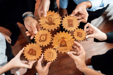 Eller, finans sembolleri sergileyen ahşap dişlilerin etrafında toplanıyor, profesyoneller arasında iş büyümesi ve strateji gelişiminde takım çalışması ve işbirliğini temsil ediyor. Dostluk