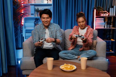 Neşeli, gülümseyen bir çiftin koltuğunda oturup oyun oynuyorlar. Oyun konsolu, hazır aperatifler ve içecekler, modern neon ışıklı, rahat oturma odasında eğlenceli bir rekabet seviyesinde. İnfobahn.