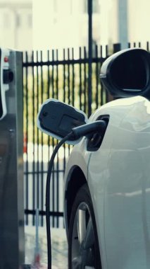 Takım elbiseli, hırslı bir işadamı elektrik kablosu ve yenilenebilir enerji enerjili bir elektrikli araçla şarj istasyonunda dururken elektrikli aracına yaslanıyor..