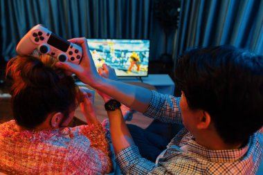 joystickli bir çift, TV ekranında dövüş oyunu oynuyor, erkek, kız arkadaşıyla yarışma seviyesini geri kazanıyor, modern evdeki neon ışıklı oturma odasında kanepeye oturuyor. İnfobahn.