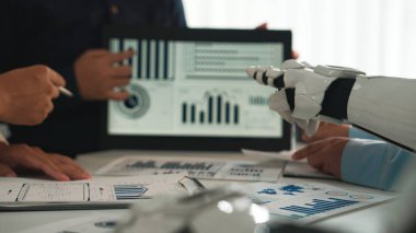 İş toplantılarında robot bir kol ve insan eli dijital ekranda veri grafiklerini analiz ederek, iş yerindeki işbirliğini ve teknolojik ilerlemeyi vurguluyor. Noogenesis