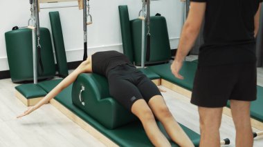 Happy Pilates eğitmeni, bir müşteriye yay namlu teçhizatı kullanarak çekirdek güçlendirme egzersizinde rehberlik ediyor. Çekici kadın bacak esnetme pozu veriyor. Vücut ve denge çalışması için, kas gücünü arttırmak için. Alışkanlık.