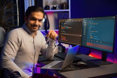 Gülümseyen akıllı bilişim geliştiricisi dizüstü bilgisayar üzerinde çalışırken kameraya bakıyor, neon ışıklı ofis odasında yazılım kodlama sistemi, uygulama programı güncellemesi yapıyor en son online web sitesi verileri. Tahmin et..