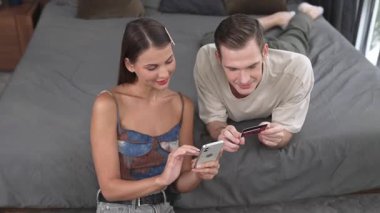 Genç çift, kredi kartıyla ödemek için internet üzerinden ödeme uygulaması ve dijital cüzdan kullanarak yatak odasında oturuyor. Mobil internet üzerinden alışveriş ve modern alımlar. Düzenle