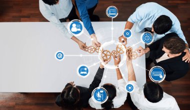 Çarklar ve teknoloji ikonlarıyla dolu bir masanın etrafında çalışan çeşitli bir ekibin hava görüşü, iş yerindeki işbirliğini, yeniliği ve iş verimliliğini gösteriyor. Dostluk