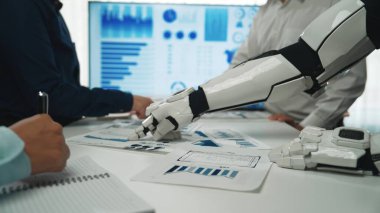 Robot bir kol modern bir ofiste iş haritaları ve belgelerle etkileşime girer. Profesyoneller, stratejik karar verme konusunda fikir alışverişinde bulunmak için veri analizinde işbirliği yaparlar.