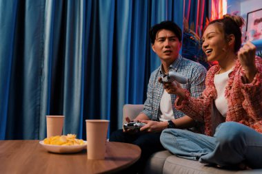 Mutlu çiftin oturduğu kanepe joysticklerle video oyunu oynuyor, meydan okuyan gülümseyen kadın, evdeki modern neon ışıklı oturma odasındaki mücadeleyi kazanmak için yumruk kaldırıyor. İnfobahn.