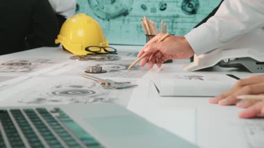 İşbirlikçi bir ekip toplantı masasında mühendislik tasarımı ve materyalleri sergiliyor, tartışmalara katılan elleri vurguluyor, araçlar ve bir dizüstü bilgisayarla birlikte. SACTR