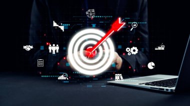 Bir işadamı, modern bir çalışma alanı ortamında çeşitli iş simgeleri ve veri grafikleriyle çevrili bir okla temsil edilen dijital bir hedefe odaklanır. Dörtgen