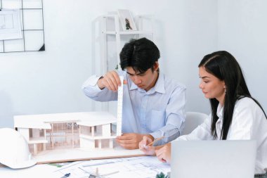 Profesyonel Asyalı erkek mimar ev modelinin uzunluğunu ölçmek için cetvel kullanırken, beyaz tenli genç meslektaşı ev modeliyle toplantı masasındaki verileri analiz etmek için dizüstü bilgisayar kullanıyor. Tertemiz..