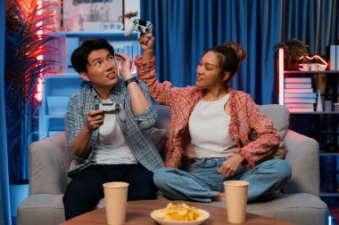 Mutlu çiftin oturduğu kanepe joysticklerle video oyunu oynuyor, meydan okuyan gülümseyen kadın, evdeki modern neon ışıklı oturma odasındaki mücadeleyi kazanmak için yumruk kaldırıyor. İnfobahn.