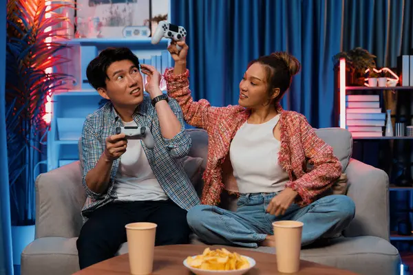 Mutlu çiftin oturduğu kanepe joysticklerle video oyunu oynuyor, meydan okuyan gülümseyen kadın, evdeki modern neon ışıklı oturma odasındaki mücadeleyi kazanmak için yumruk kaldırıyor. İnfobahn.