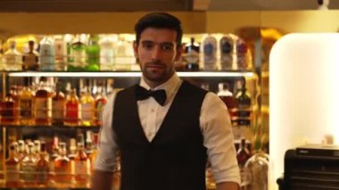 İyi görünümlü, gülümseyen, bardaki lüks içki tezgahında cam silen barmen profili. Bar gece kulübünde sıcak karşılayan profesyonel barmen kostümünü poz vermek için kameraya bakıyor. Vinosity.