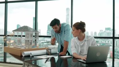 Akıllı mimar mühendis ev modelini incelerken meslektaşı da laptop analiz verilerini kullanıyor. Modern ofisteki ev modeli inşaatında profesyonel tasarımcı takımı birlikte çalışıyor. İzleme