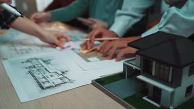 Takım üyeleri masadaki planlar ve mimari modelleri inceleyerek, modern ofis ortamında takım çalışması ve yaratıcılığı sergileyerek işbirliği yaparlar. SACTR