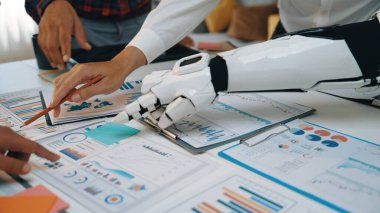 Bir robot kol, basılı raporlarda veri analizi yapar, canlı bir ofis ortamında insan ve makine işbirliğinin bir karışımını gösterir. Noogenesis