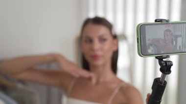 Genç sosyal medyum kadın moda videosu yapıyor. Blogcu kameraya selfie çubuğu ve ışık halkası kullanarak gülümserken, aynı zamanda ikna edici çevrimiçi kıyafetlerin izleyicilere veya takipçilere vlog satmasını sağlıyor. Düzenle