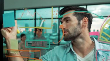 Akıllı proje yöneticisi cam duvara yapışkan notlar ve planlama stratejisi için zihin haritası yazarken, aynı zamanda çok kültürlü iş adamları da şehir manzaralı iş planlarını tartışıyorlar. İzleme